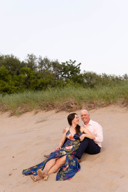 woman in blue floral long dress sits on sand in front of husband in a suit