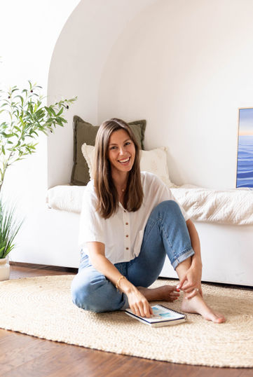 woman in white shirt and blue jeans sitting on floor with sketchpad smiling at camera