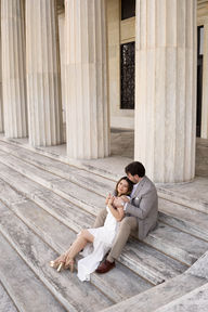 woman in white dress and man in gray suit sit leaning into each other on marble steps of Buffalo History Museum