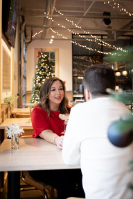 woman reaches across a table to hold her fiances hand, string lights hang from the ceiling