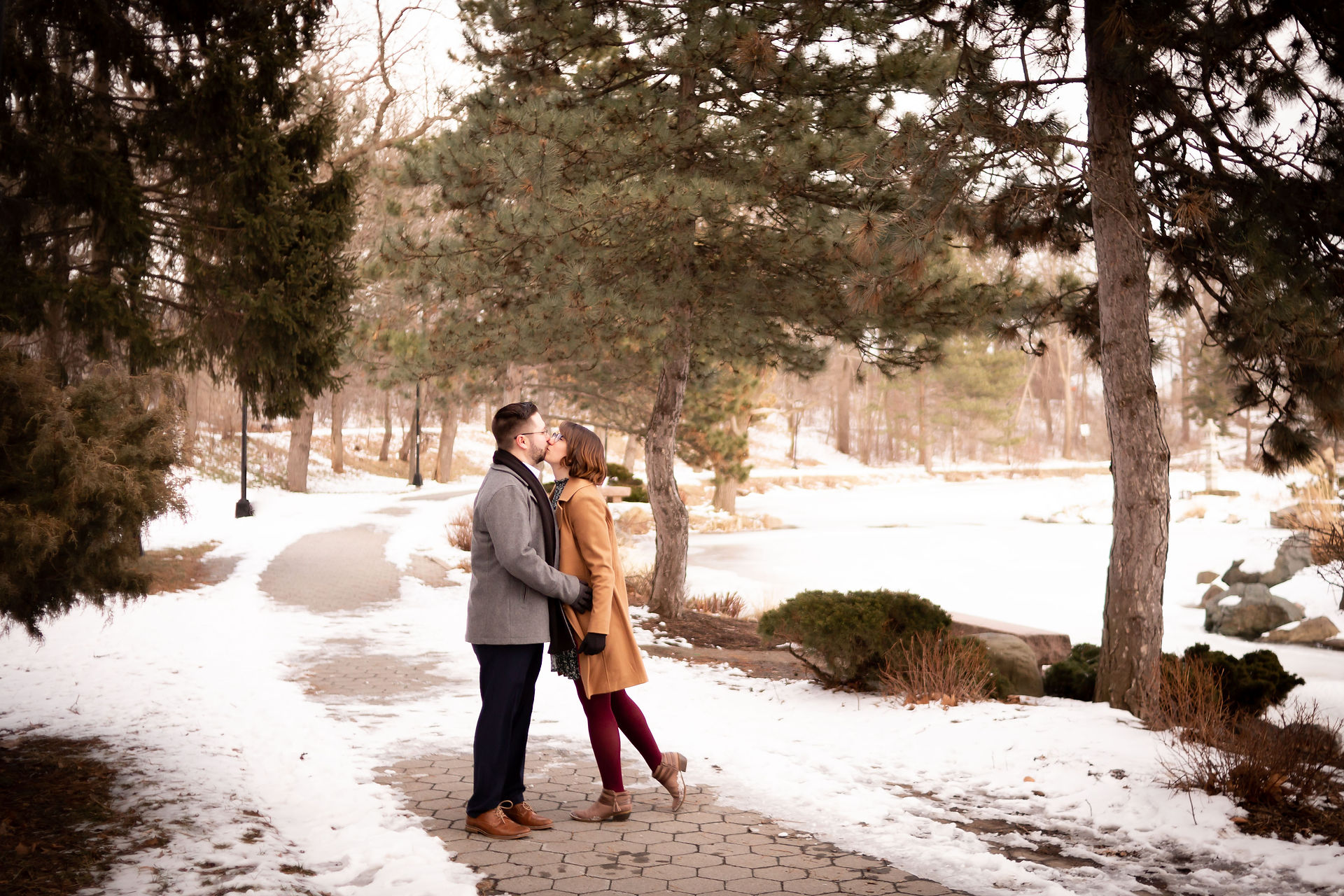 snow lined path with trees and bushes and a couple standing in the middle kissing