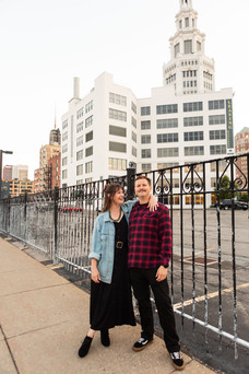 couple stands on city street with man looking at the camera and woman with her arm around the man's shoulder looking at him
