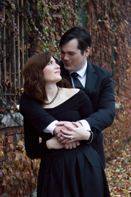 woman in black dress stands in front of man in black suit who has his arms around her. they look at each other