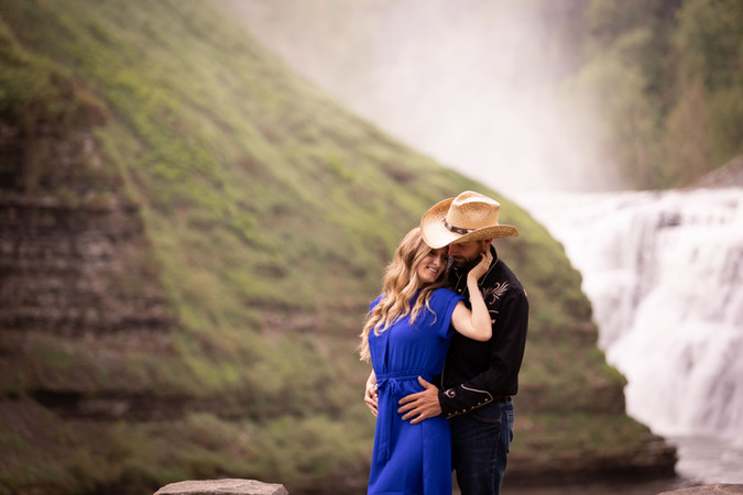 woman stands with her back to a man and her hand up on his cheek. he's wearing a cowboy hat. waterfall is in background.