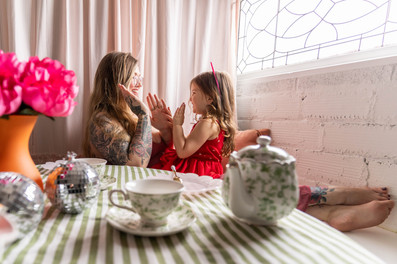 mom and daughter sit at tea party with floral tea set, green and white striped tablecloth and pink curtains playing patty cake