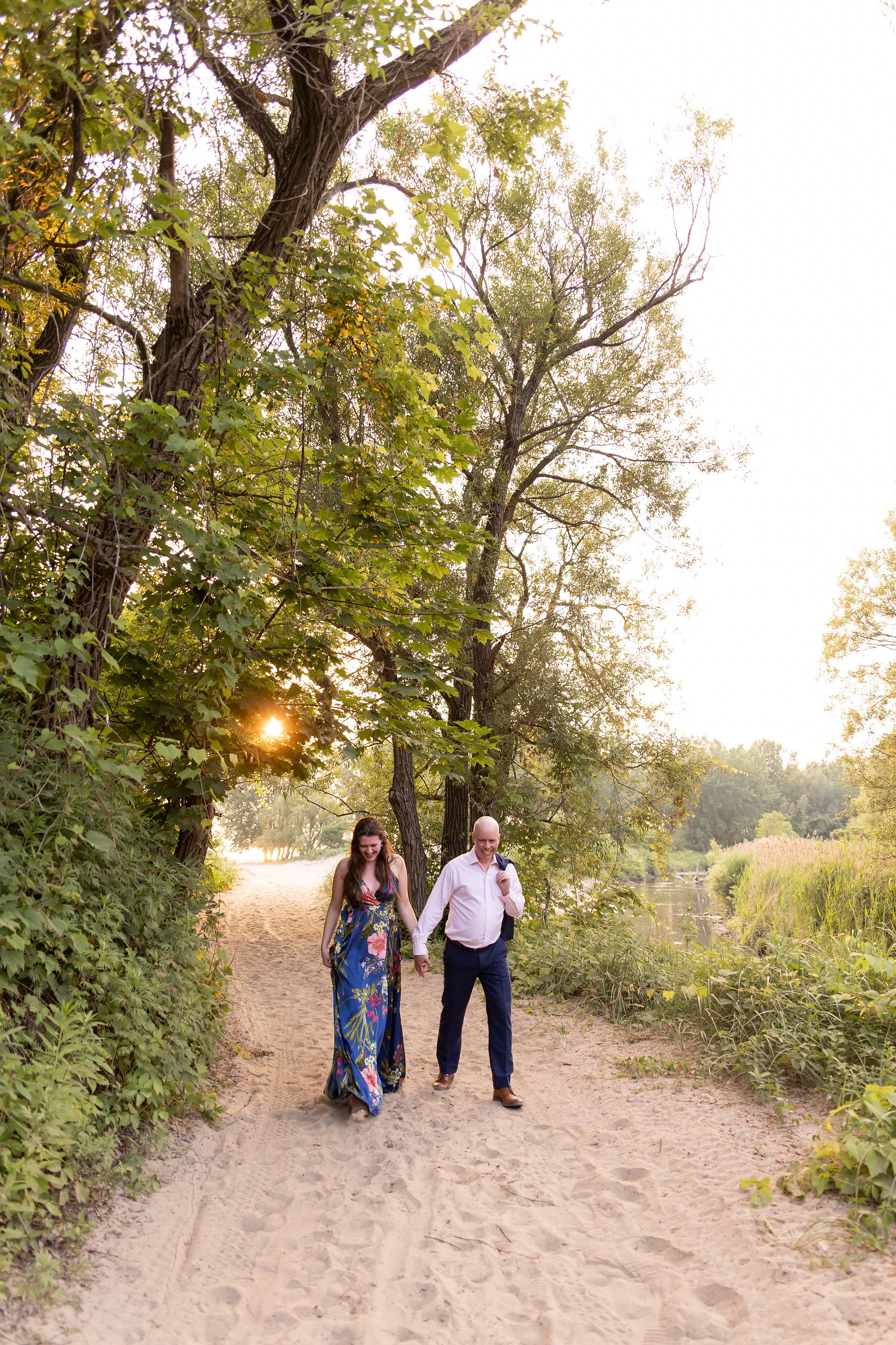 couple walks hand in hand on sand through trees with sun shining through the trees