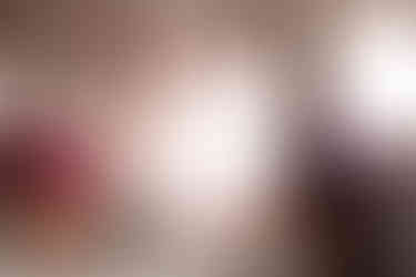 pink flowers in an orange vase sit next to a green and white tea kettle on a table with a woman blurred out in the background in purple lingerie