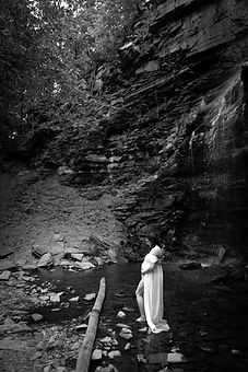 woman with a baby bump stands in front of a waterfall in Buffalo with a white dress on that reveals her pregnant stomach
