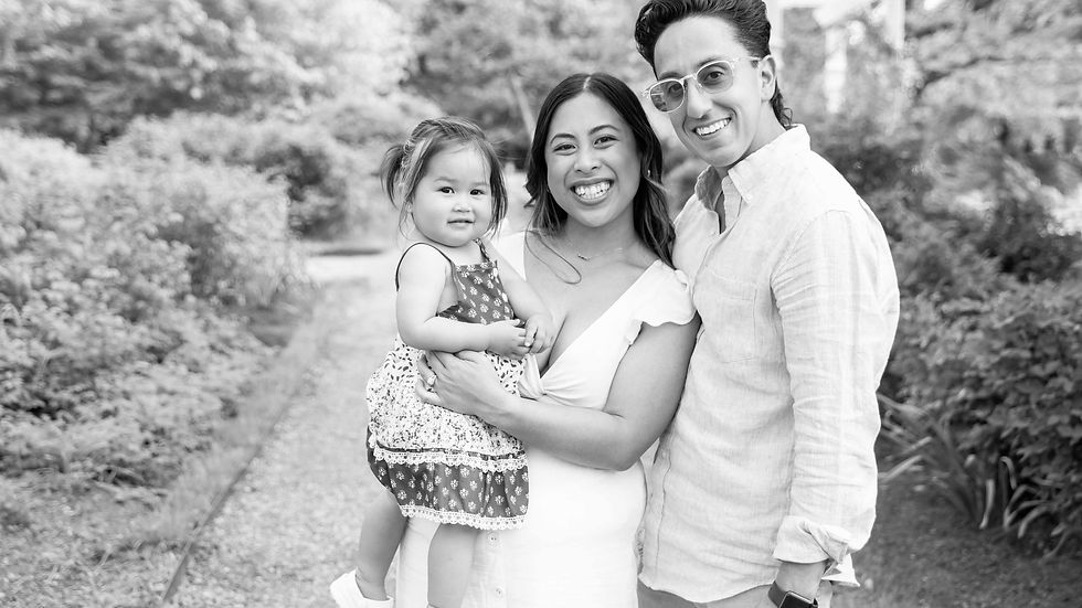 A smiling family poses in a garden, woman holding a toddler in a patterned dress. Lush greenery in the background, black and white photo.