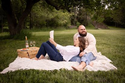 couple sits on a blanket in a park, woman leans into man while she reads her book, a picnic basket and two glasses of white wine in background