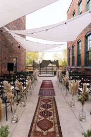 looking down the aisle of a wedding ceremony with vintage rugs lining walkway, pampass grass in glass chairs at the end of each row of seats and large white shade covers strung between two brick buildings