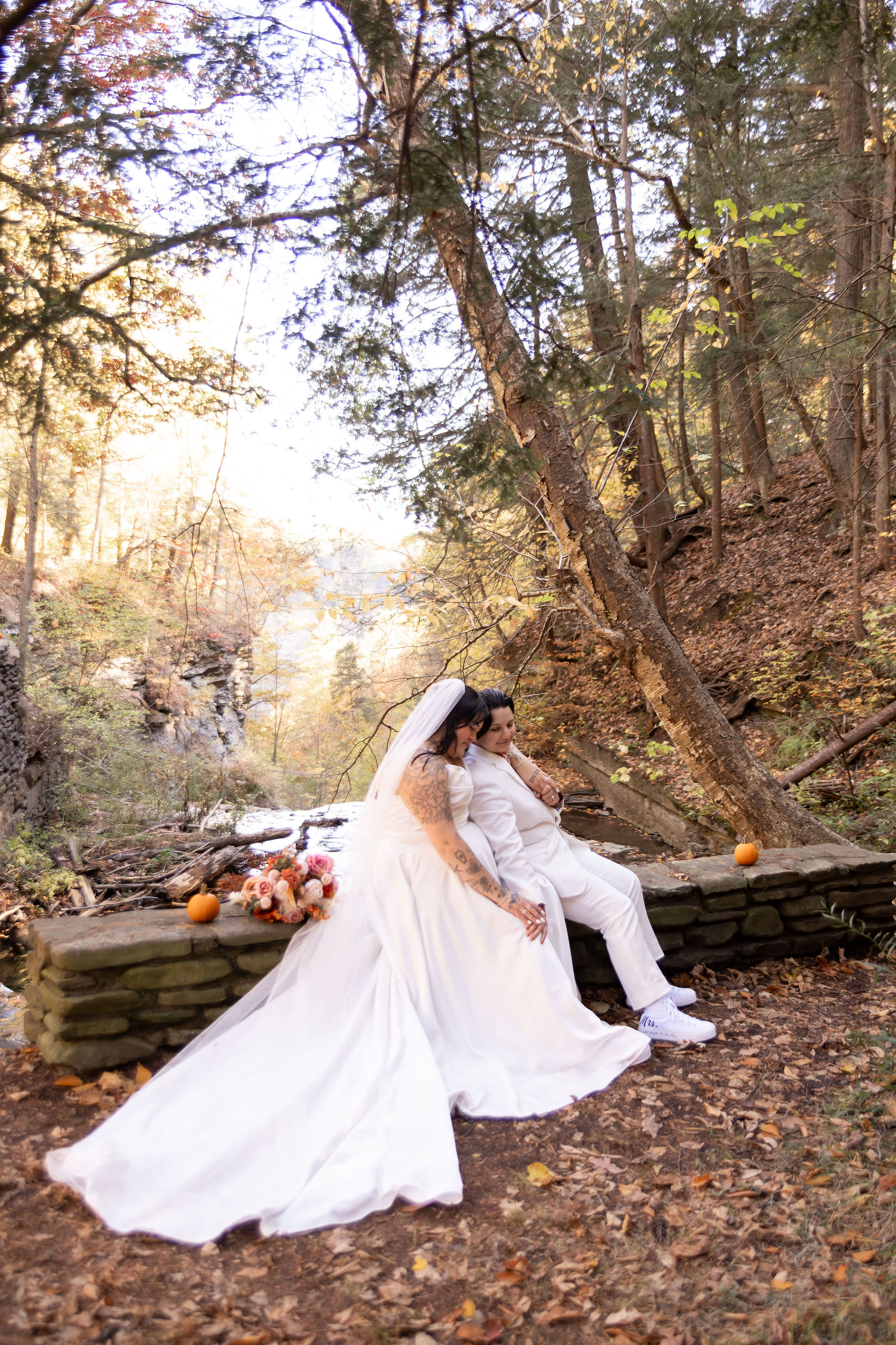 bride in white dress sits behind her bride in white suit on cliff edge overlooking Letchworth canyon