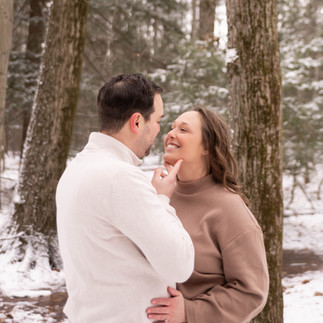 man in white sweater holds chin of fiance in brown sweater, they stand amongst snow covered trees