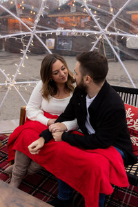 man and woman chat on a couch inside a winter igloo with a red blanket on their laps