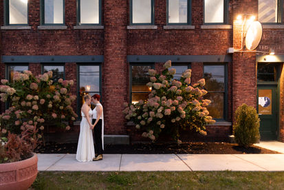 two brides stand forehead to forehead in front of waxlight bar brick building in Buffalo