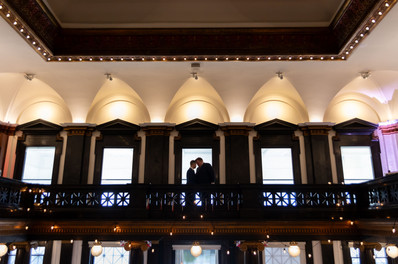 silhouette of two grooms standing forehead to forehead in front of row of arched windows