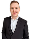 A man and dark brown hair smiling in a black suit