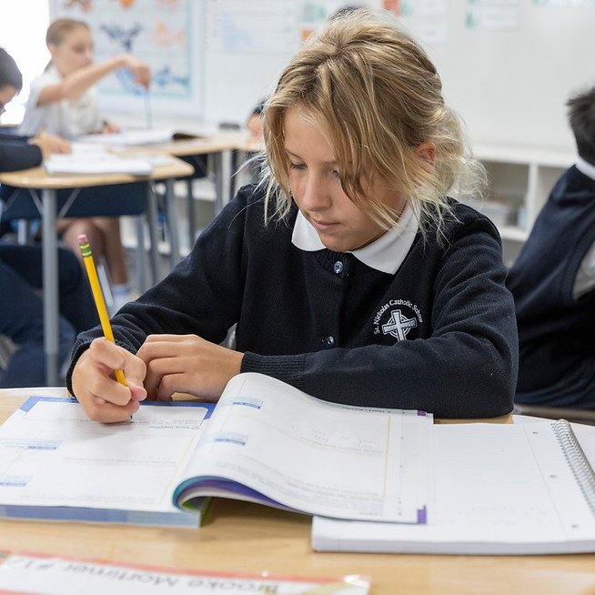 This image links to our giving portal. It depicts a girl in 5th grade writing in a workbook at Saint Nicholas Catholic School in Los Altos California.