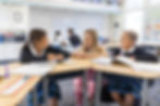 A teacher kneeling at a desk and talking with two fourth grade girls who are working on classwork at Saint Nicholas Catholic School