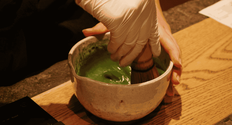 a person's hand mixing a matcha mix with a matcha bamboo whisk