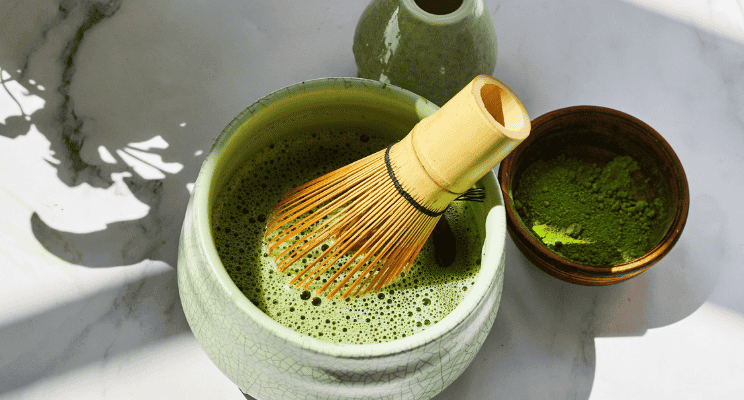 matcha mixture in a bow with a matcha bamboo whisk in it beside a bowl of matcha powder, and water