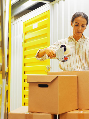 portrait-of-young-woman-packing-boxes-with-tape-gun-while-standing-by-self-storage-unit-co