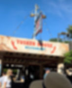Tusker House Restaurant entrance sign under blue sky, with visitors walking below and string lights overhead.