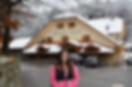 Smiling woman in a pink jacket stands before a snowy lodge labeled Domačija Vodnik, with parked cars and winter trees.