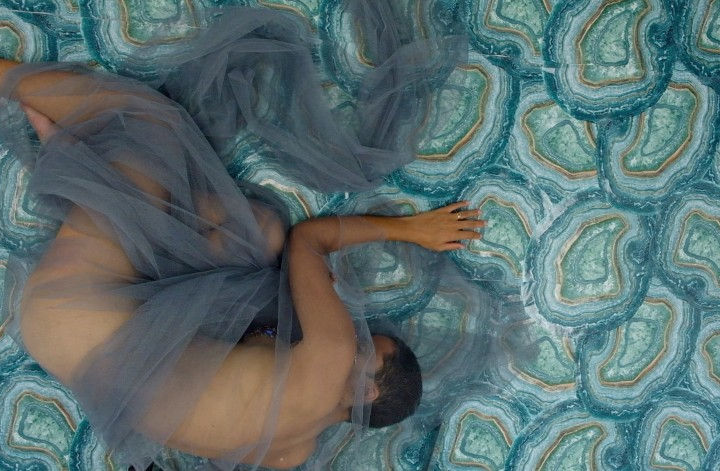 A naked light-skinned Black person lies on a blue-agate floor. They are wrapped in blue tulle material.