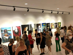 Sanat galerisinde, duvarda resimler asılı ve insanlar sergiyi geziyor. Kalabalık ortam, samimi bir atmosfer sunuyor.