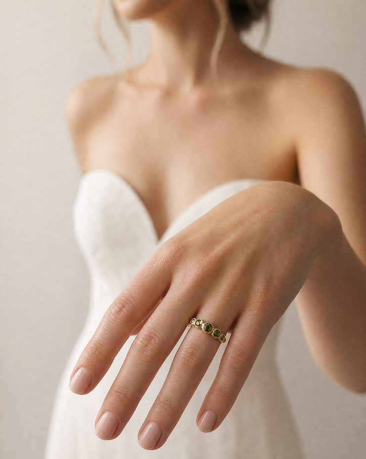 Close-up of a woman in a wedding dress showing a gold ring with green diamonds against a soft beige background.