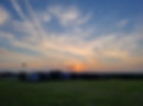 Pic description: view of a sunset field. Evening light, dark green grass in forefront, trucks and vans lined up, some with flags, in shadowy darkness, behind and above is an orange and blue swirly sky with fluffy white clouds.