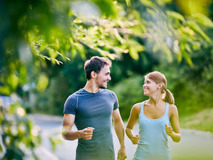 A man and woman jog on a leafy path, smiling at each other. They wear athletic gear, and the background is lush and green, creating a lively mood.