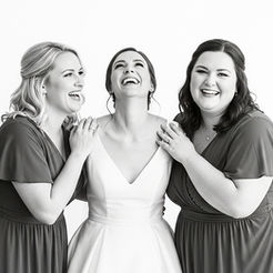 Black-and-white photo capturing three women—one bride in a white dress and two bridesmaids in matching dark dresses—sharing laughter and happiness at a wedding.