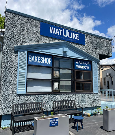 The exterior of the building. Blue building with lots of windows. Has two benches for outdoor seating and small tables. Plus beautiful planters