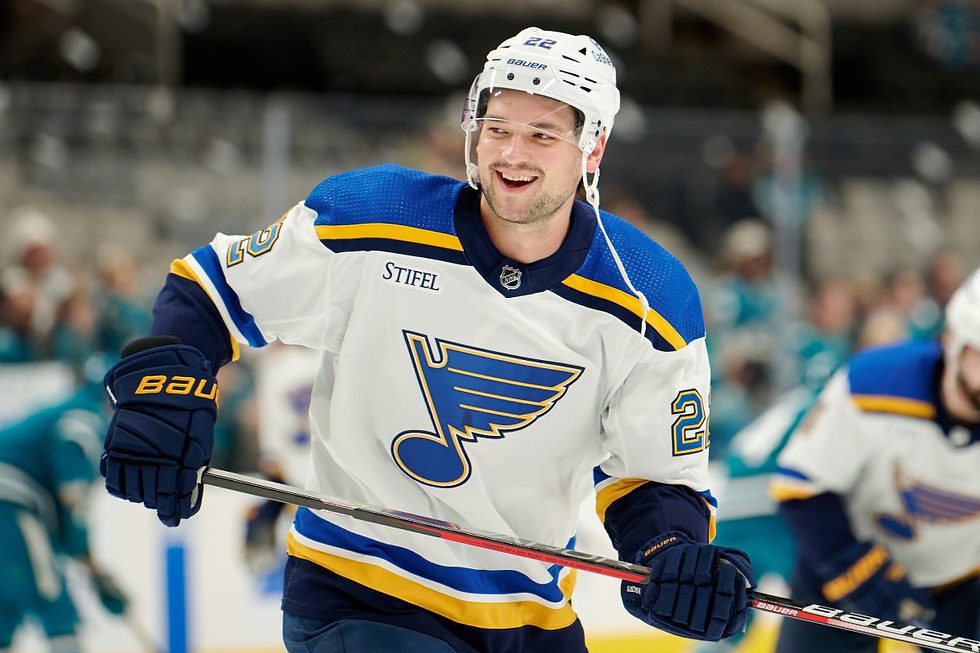 Logan Brown holds his hockey stick and smiles at someone off screen while wearing a white St. Louis Blues jersey with the number 22 on the shoulders.