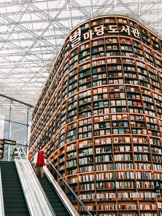 A towering bookshelf inside Starfield Library in Seoul, South Korea, with a person in a red jacket ascending an escalator beneath the glass ceiling.