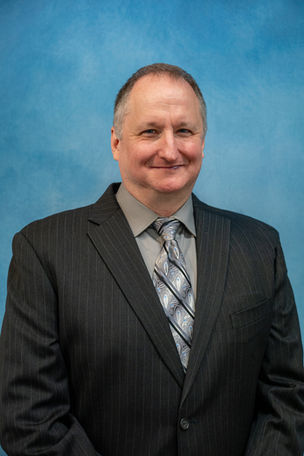 Corporate headshot of a businessman in a dark suit and patterned tie against a blue studio background.