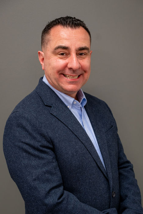 Business headshot of a man in a navy blazer and open collar shirt against a warm brown backdrop.