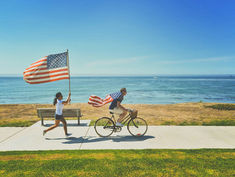 Man cycling with American flag cape, followed by girl running holding flag. Ocean and blue sky in background, creating a joyful scene.