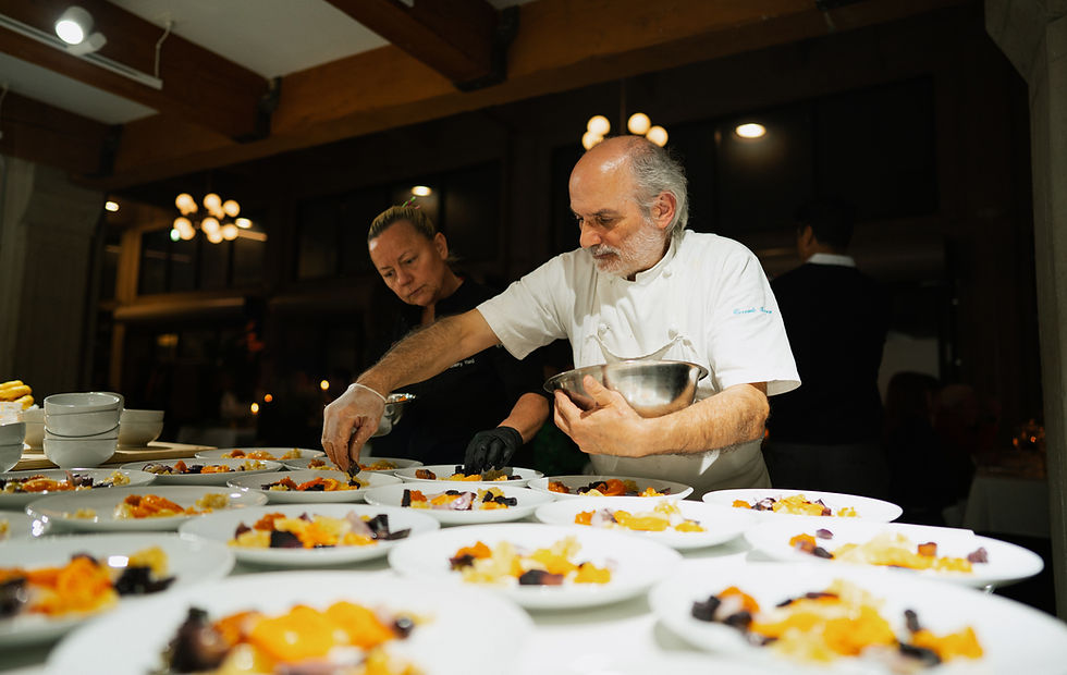 Two chefs plate colorful dishes in a dim restaurant kitchen, one holding a metal bowl under warm lights.