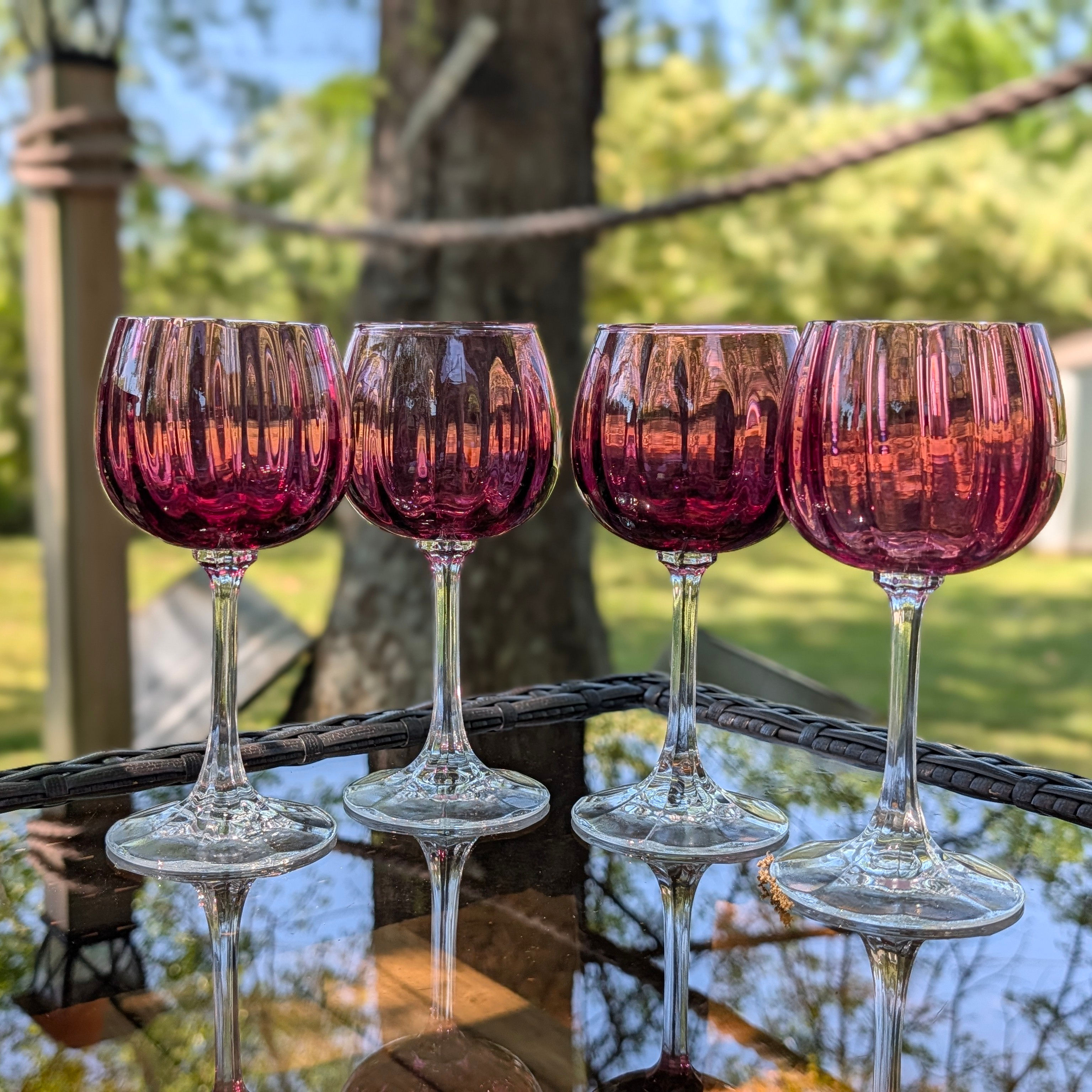 set of 4 cranberry glass wine glasses on glass table in outdoor setting with lush green landscape background on a sunny day