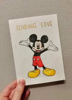 A hand holds a card with Mickey Mouse printed on the front with the words "Sending love"