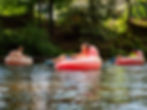 Three kids float on pink inflatables in a river, surrounded by lush green trees. The mood is relaxed and playful.
