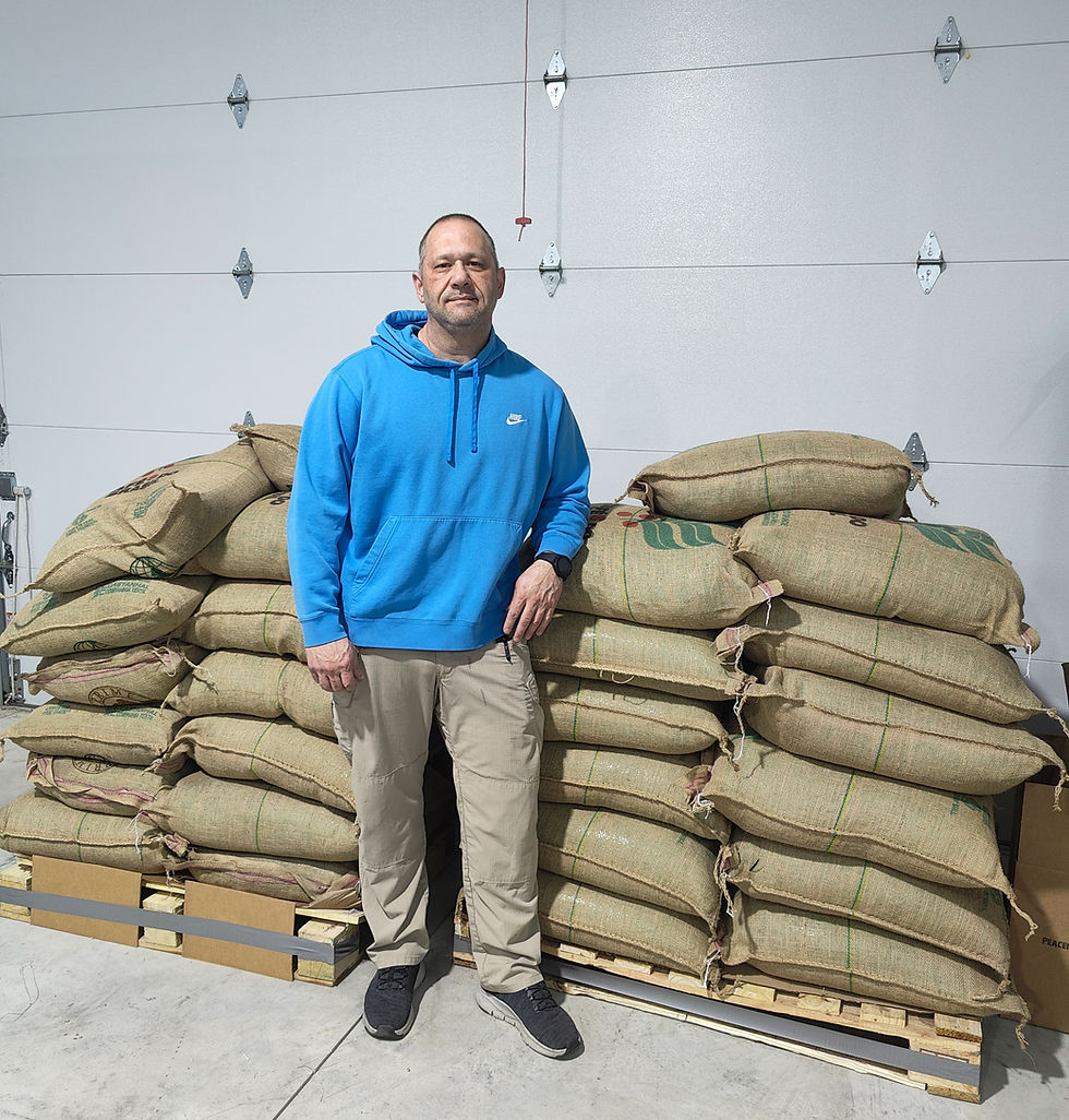 Chris Morris (Founder/CEO) is standing next to bags of raw green coffee beans in a warehouse.