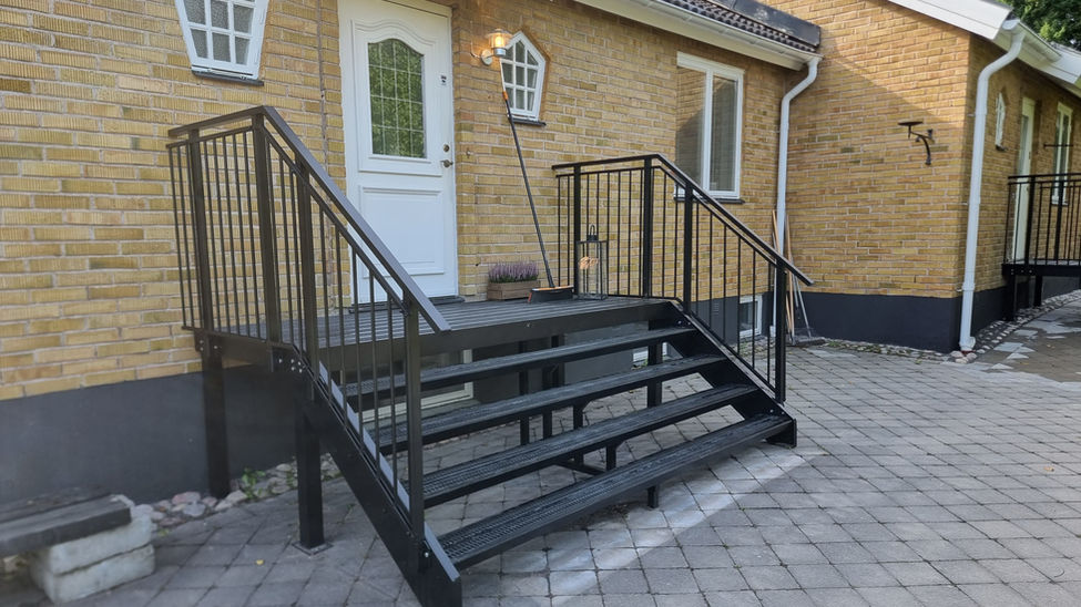 Bred, svartlackerad entretrappa i aluminium. Trappan har barnsäkra räcken och är monterad på ett hus av tegel.