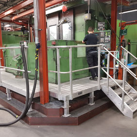 Arbetsplattform i aluminium med trappa i vardera ände. Den står uppställd vid en grön CNC-maskin i en fabrik.