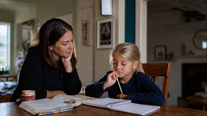 Parent listening patiently as a daughter pauses over schoolwork at a dining table.