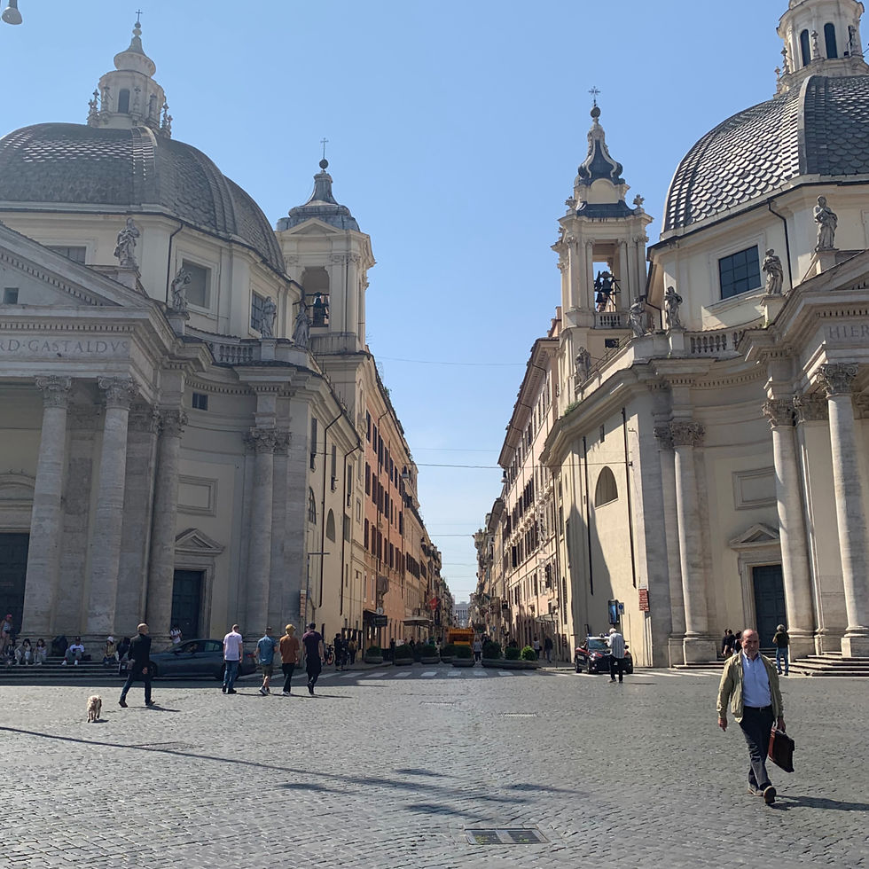 People walk across a cobbled piazza between two domed churches under a clear blue sky. A man walks a dog. Urban architecture is prominent. Rome's Piazza del Popolo.