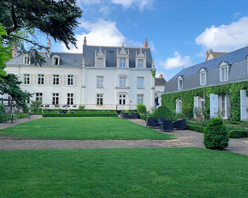 Elegant white mansion and ivy-covered wing overlook a manicured green garden under a bright blue sky with fluffy clouds.